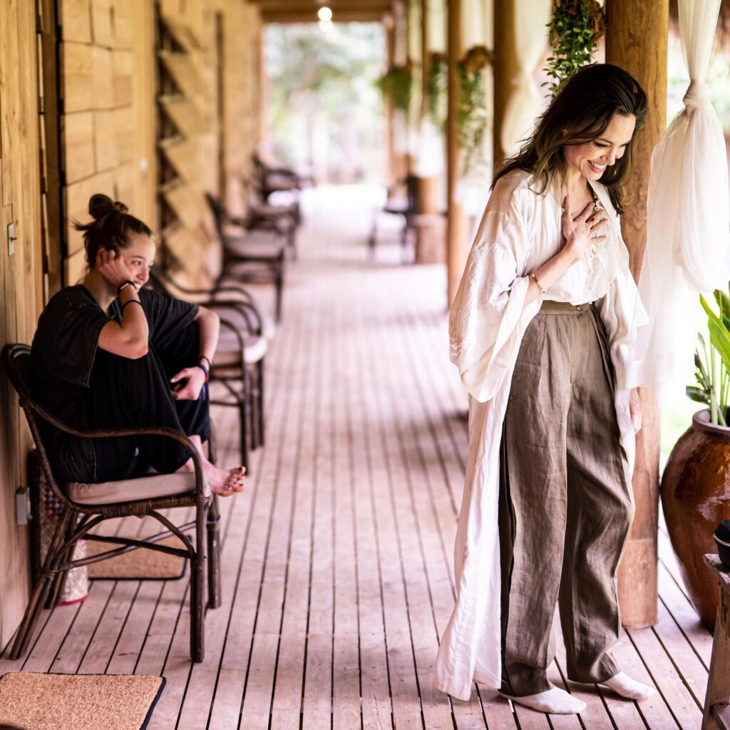 Angelina Jolie smiles softly while wearing a white robe and linen pants, standing on a wooden porch. A pensive young woman sits nearby.