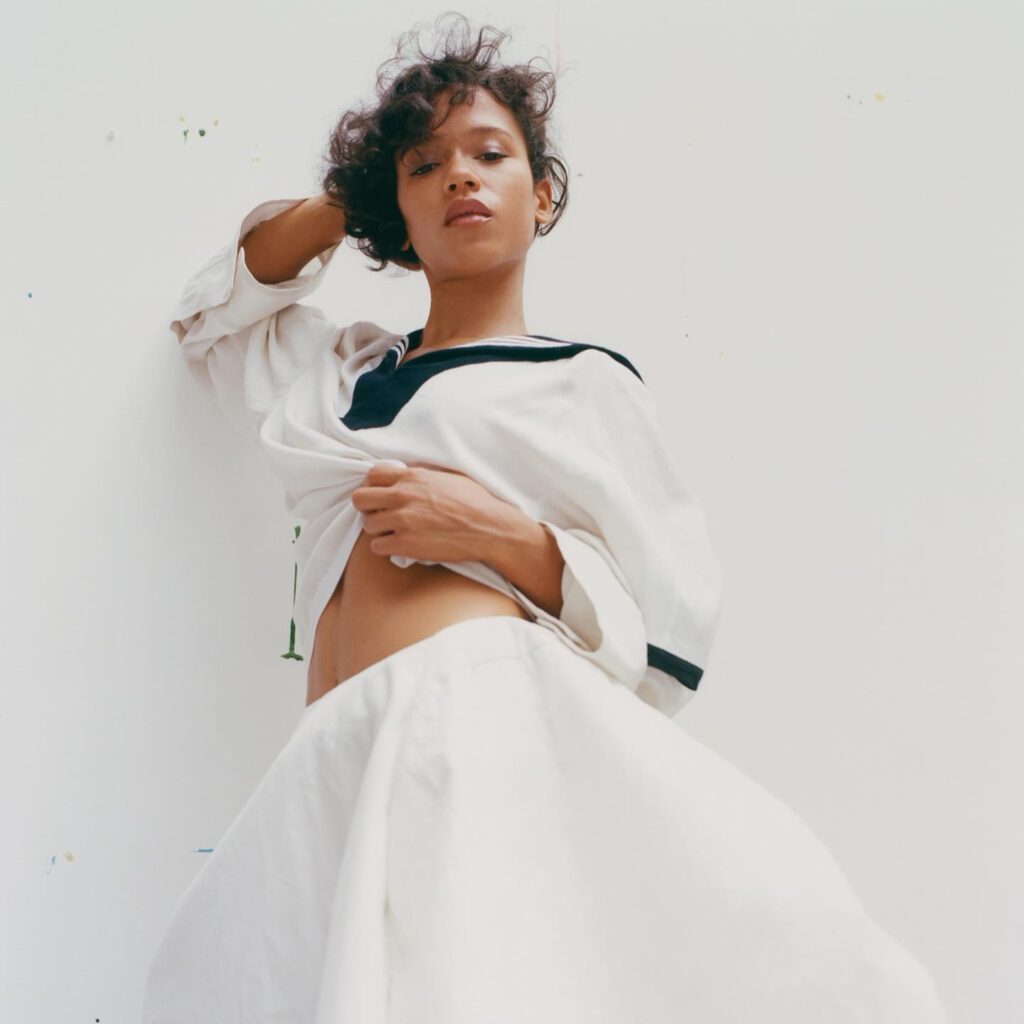 A young woman with curly hair poses confidently, wearing a white top with a black collar, showcasing her midriff against a plain background.