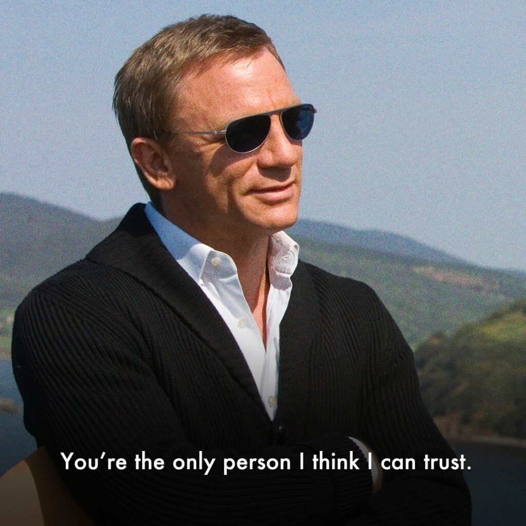 Daniel Craig, dressed in a sleek black suit with gloves and sunglasses, stands poised outdoors with a serious expression.

