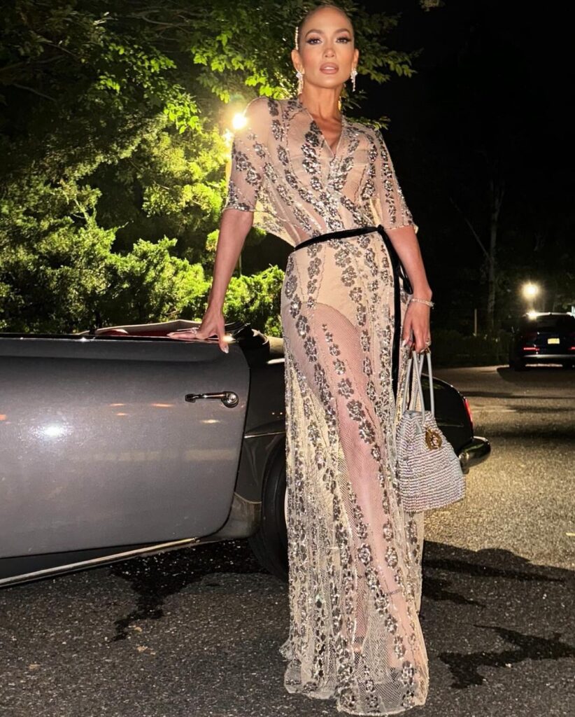 Jennifer Lopez stands elegantly beside a vintage car, wearing a stunning sheer beige gown adorned with intricate embellishments. She carries a small handbag and poses confidently under soft lighting in a nighttime setting.