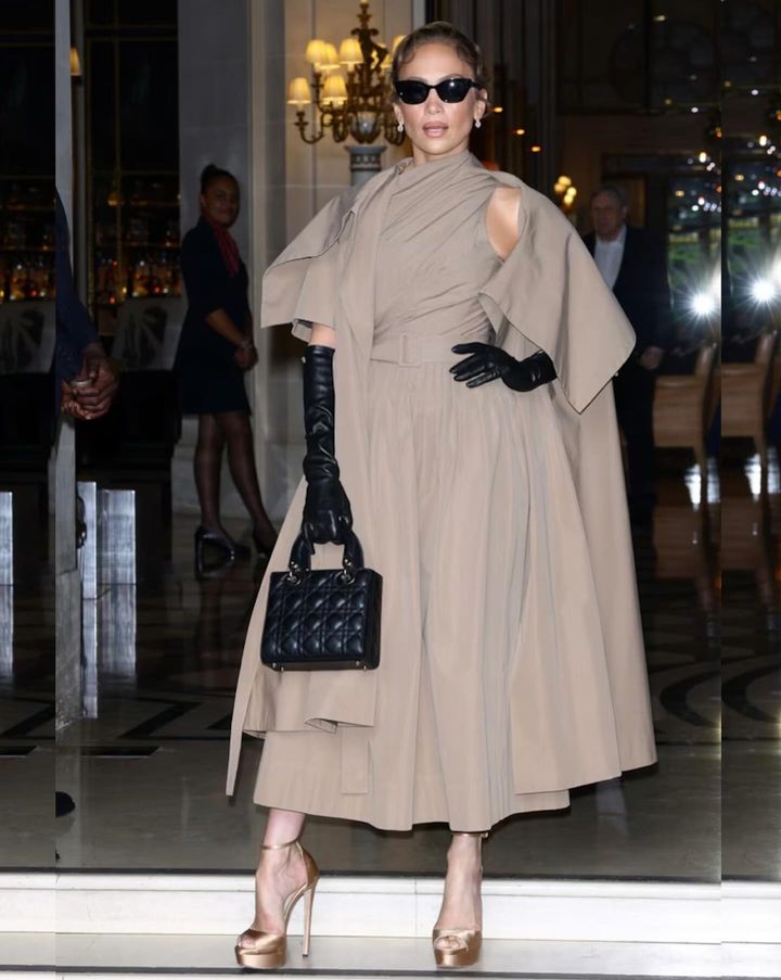 Jennifer Lopez poses stylishly in a beige couture dress with dramatic sleeves, black gloves, strappy heels, and sunglasses, holding a black handbag, against an ornate backdrop.