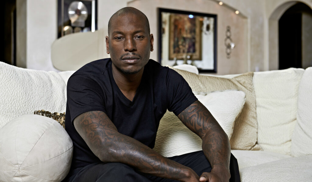 Tyrese Gibson sits confidently on a couch, wearing a black shirt, showcasing his tattoos with a serious expression.