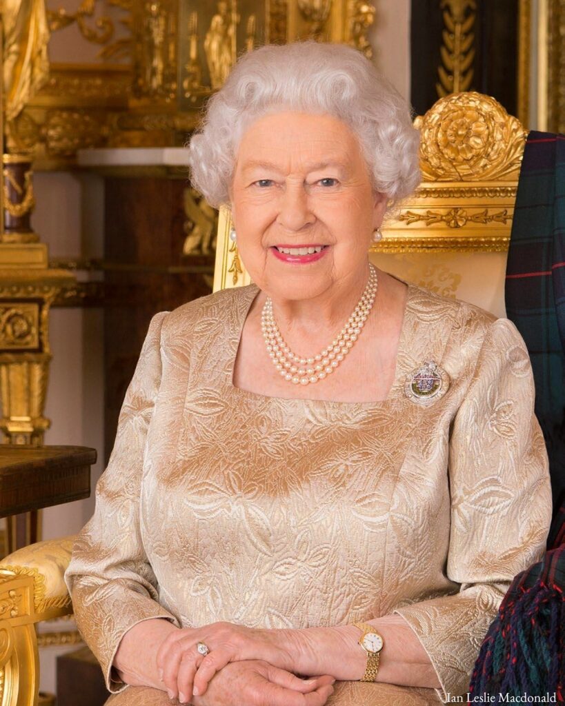 Queen Elizabeth II seated in a golden chair, wearing a pearl necklace and a gold brocade dress, smiling warmly at the camera.