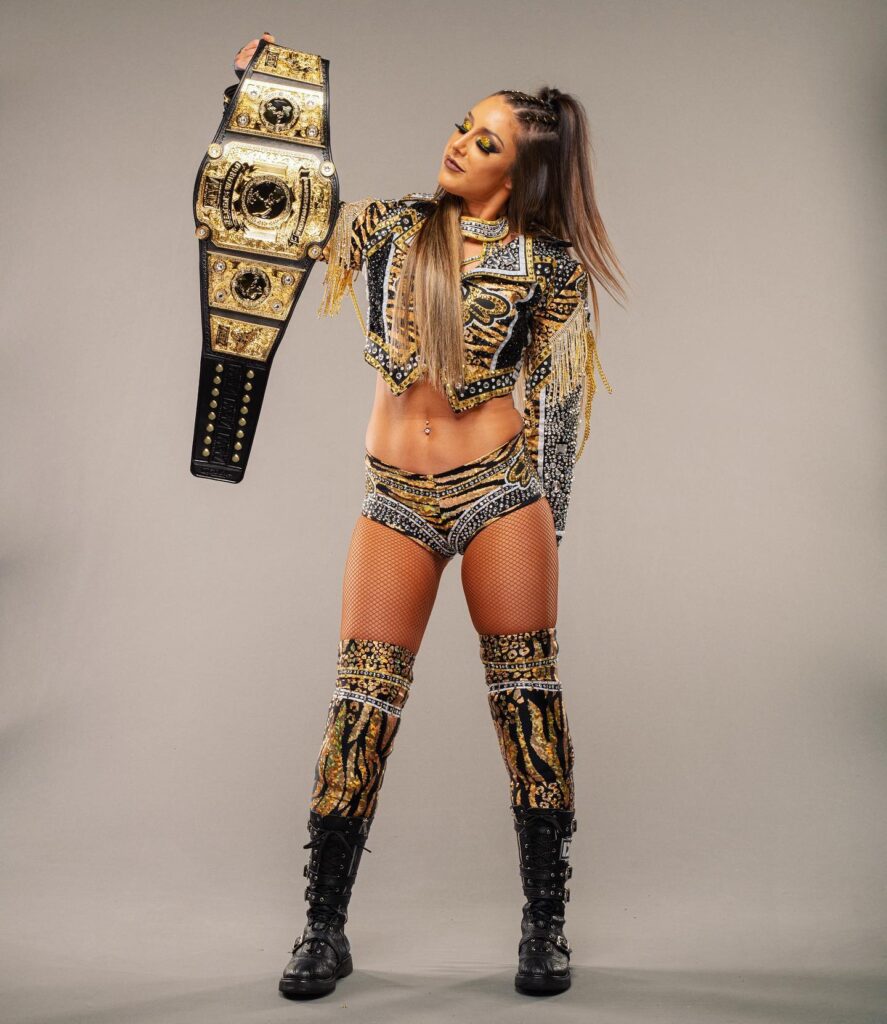 Britt Baker poses proudly holding her championship belt, wearing a dazzling gold and black wrestling outfit with intricate designs, showcasing her confident demeanor.

