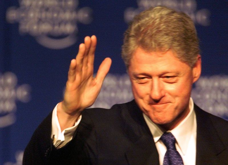 Bill Clinton waving and smiling while attending an event, wearing a suit and tie, with the World Economic Forum logo blurred in the background.