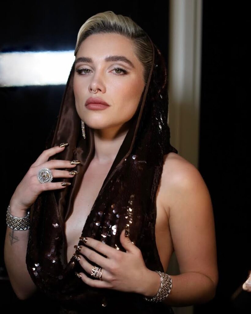 Florence Pugh poses elegantly in a sequined gown with a hood, showcasing statement jewelry and bold nails.