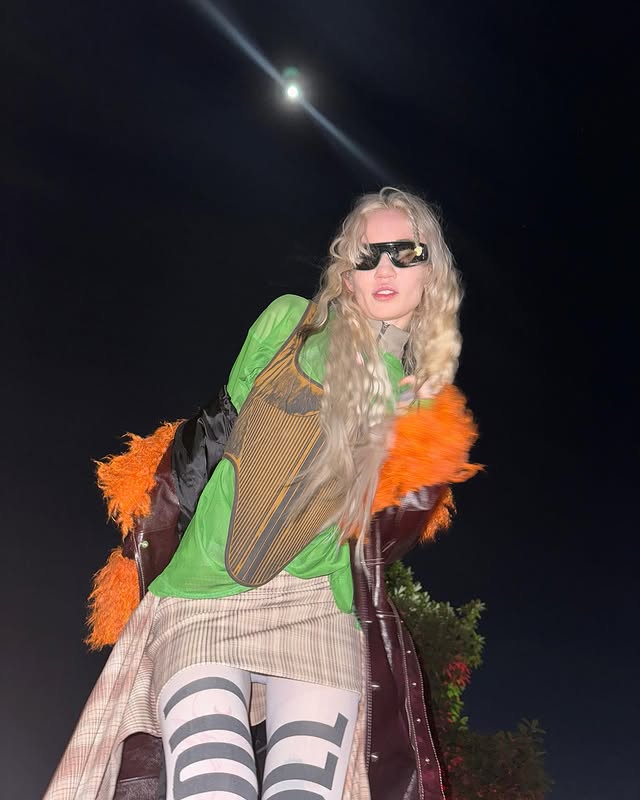 Grimes poses confidently at night, wearing a vibrant green outfit with orange fur accents, large sunglasses, and patterned stockings, against a dark sky illuminated by a full moon. Her long hair cascades down as she strikes a bold stance.