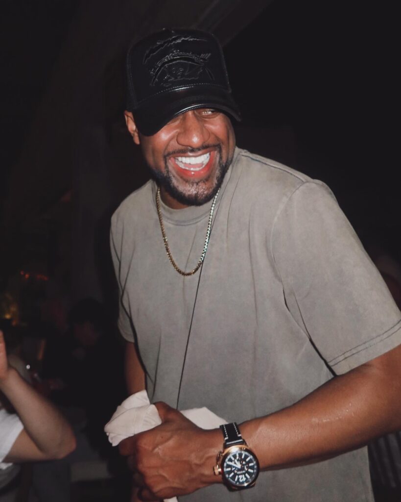 Jaleel White smiles joyfully while holding a cloth napkin in a dimly lit social setting.