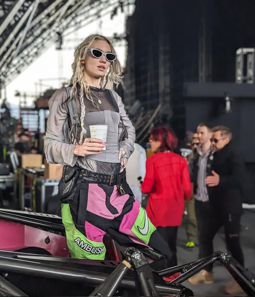 Grimes stands confidently at a music event, wearing a stylish outfit with colorful shorts, holding a coffee cup, and showcasing her unique fashion sense.