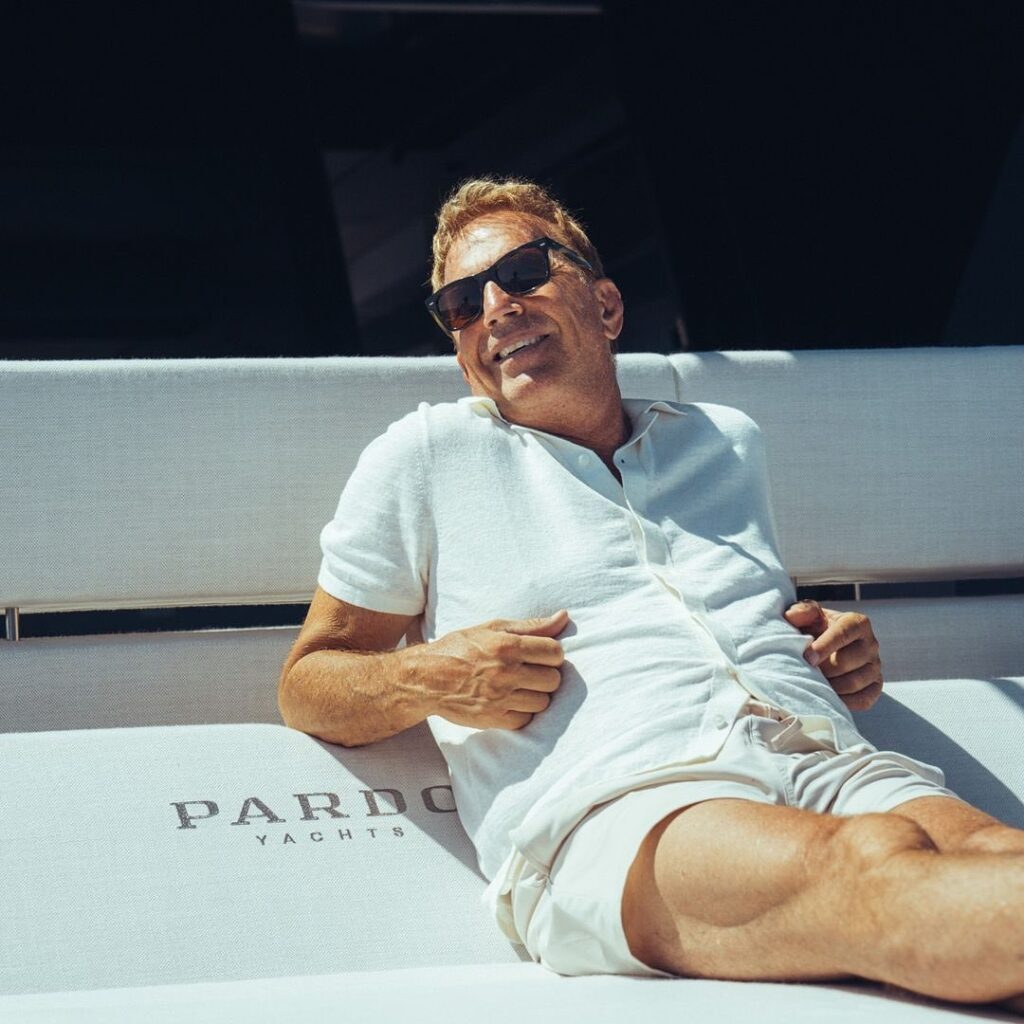 A smiling man with sunglasses relaxes on a yacht, wearing a light shirt and shorts, with the words "Pardo Yachts" visible on the cushion.

