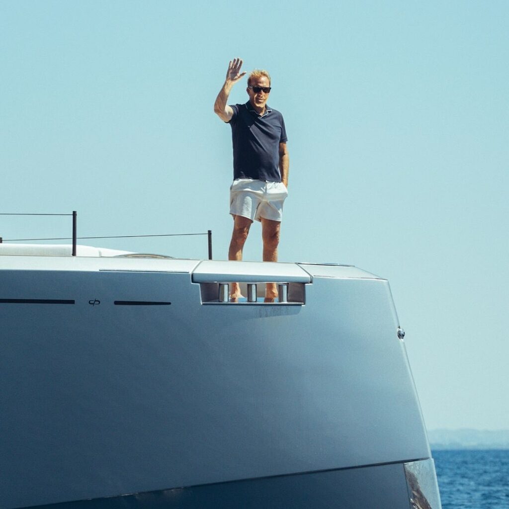 A man with sunglasses waves from the bow of a sleek yacht against a clear blue sky.