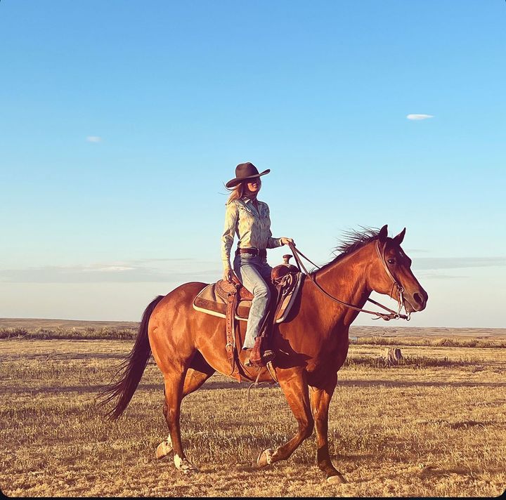 A confident woman rides a brown horse across a wide, open field under a clear blue sky, dressed in a button-up shirt, jeans, and a cowboy hat.