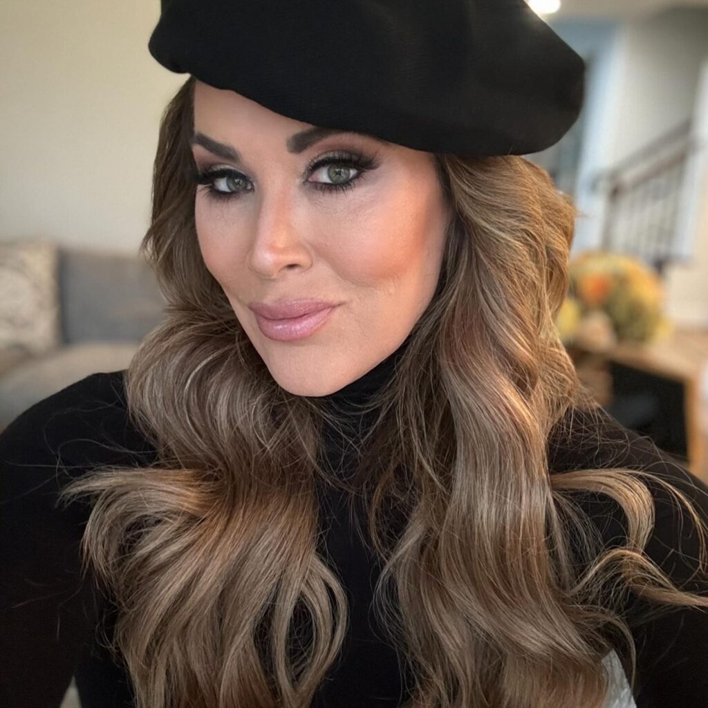 A woman with long, wavy hair and a black beret poses for a close-up selfie, featuring dramatic makeup and a black turtleneck.