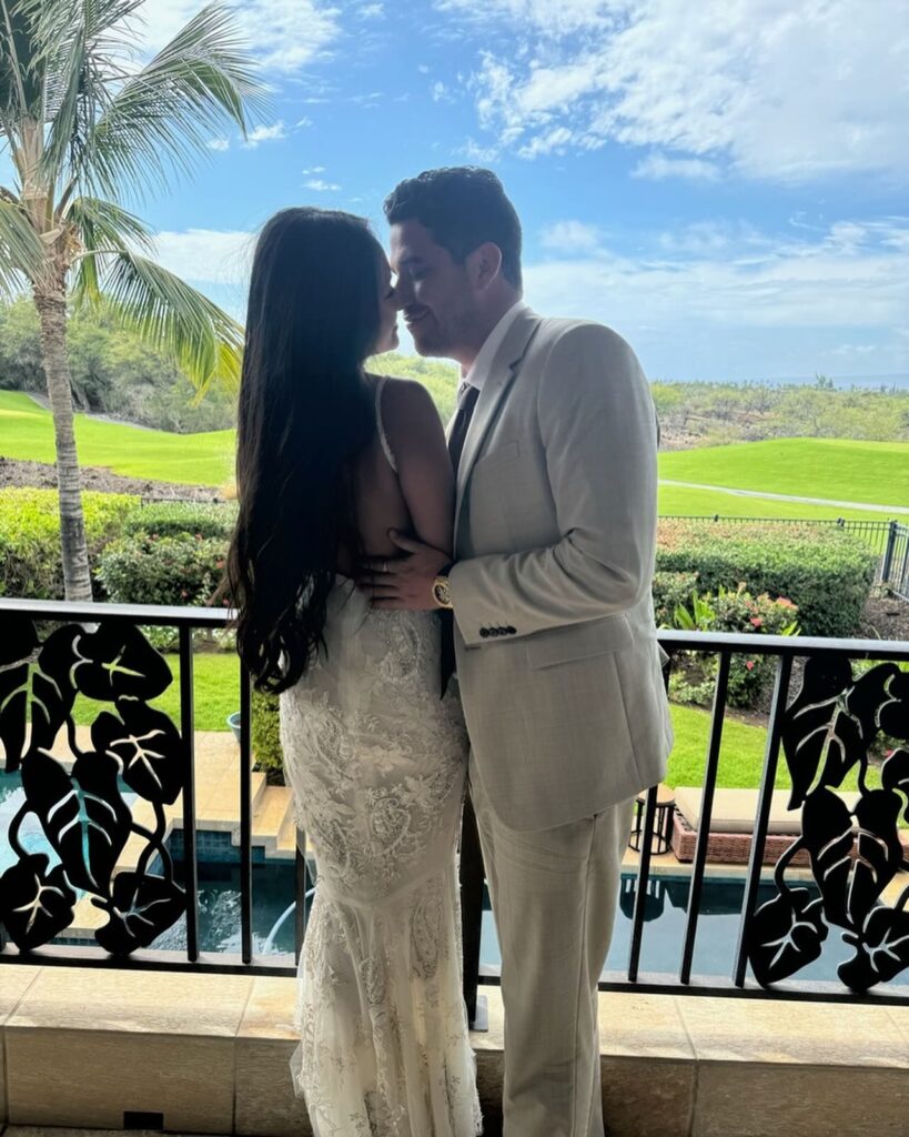 A couple shares a romantic moment, leaning in for a kiss on a balcony, dressed elegantly against a backdrop of greenery and a clear blue sky.