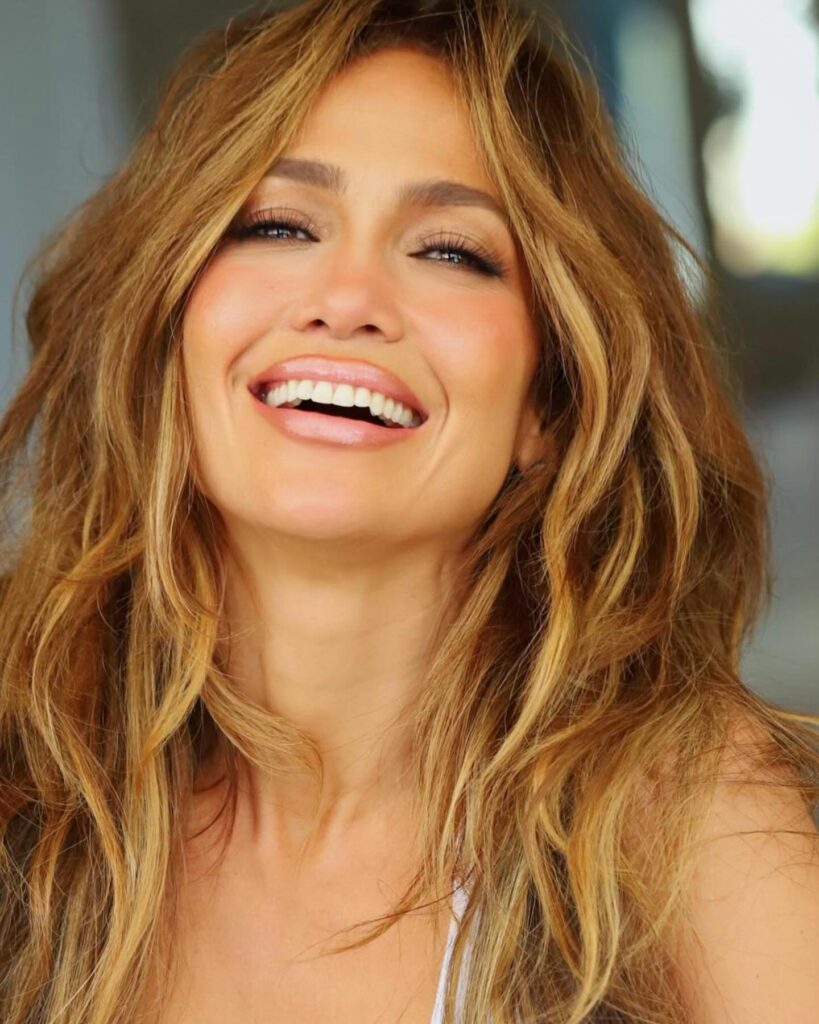 Close-up of Jennifer Lopez smiling radiantly, showcasing her wavy, sun-kissed hair and glowing skin.