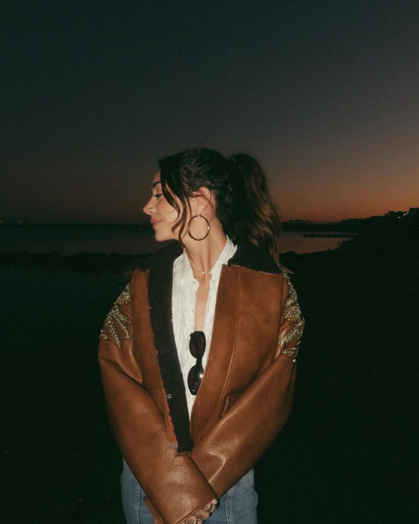 Brianna Chickenfry standing by the water at sunset, wearing a brown jacket and white shirt, with her hair tied back.