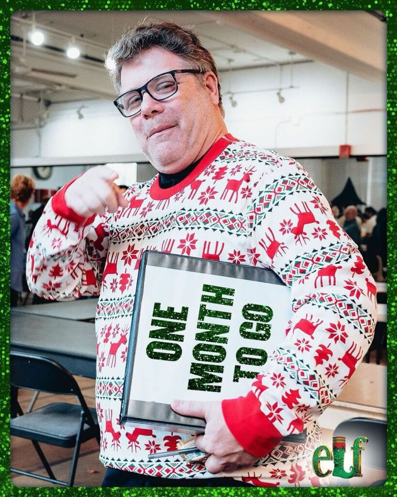 Sean Astin smiles and points to a sign reading "ONE MONTH TO GO" while wearing a festive Christmas sweater adorned with reindeer, surrounded by a cheerful holiday atmosphere.