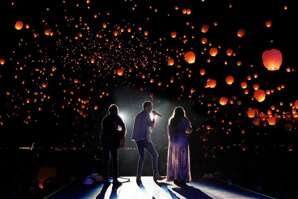 Three performers stand on stage silhouetted against a night sky filled with floating orange lanterns, creating a magical atmosphere.