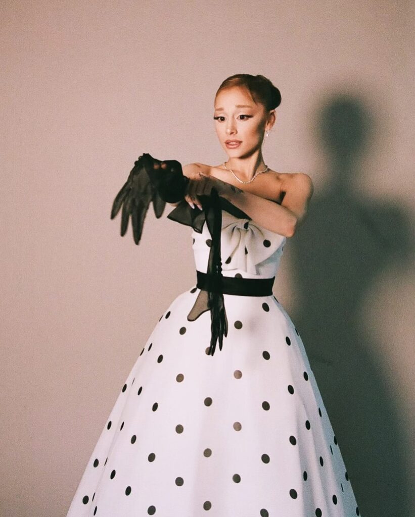 A young woman in a strapless, polka-dot gown with black accents gracefully poses while extending her arms, as if showcasing delicate black gloves. She has an elegant hairstyle and sparkles with jewelry against a softly lit background.