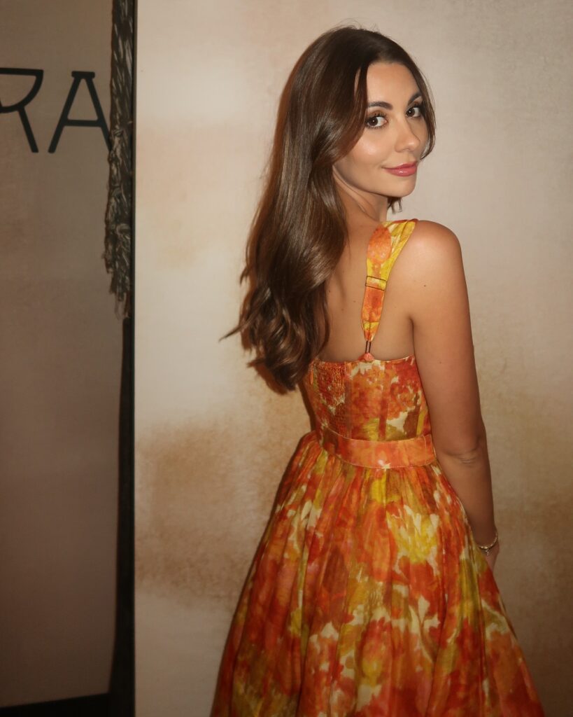 Kelley Flanagan poses in a vibrant floral dress, looking over her shoulder against a textured background.