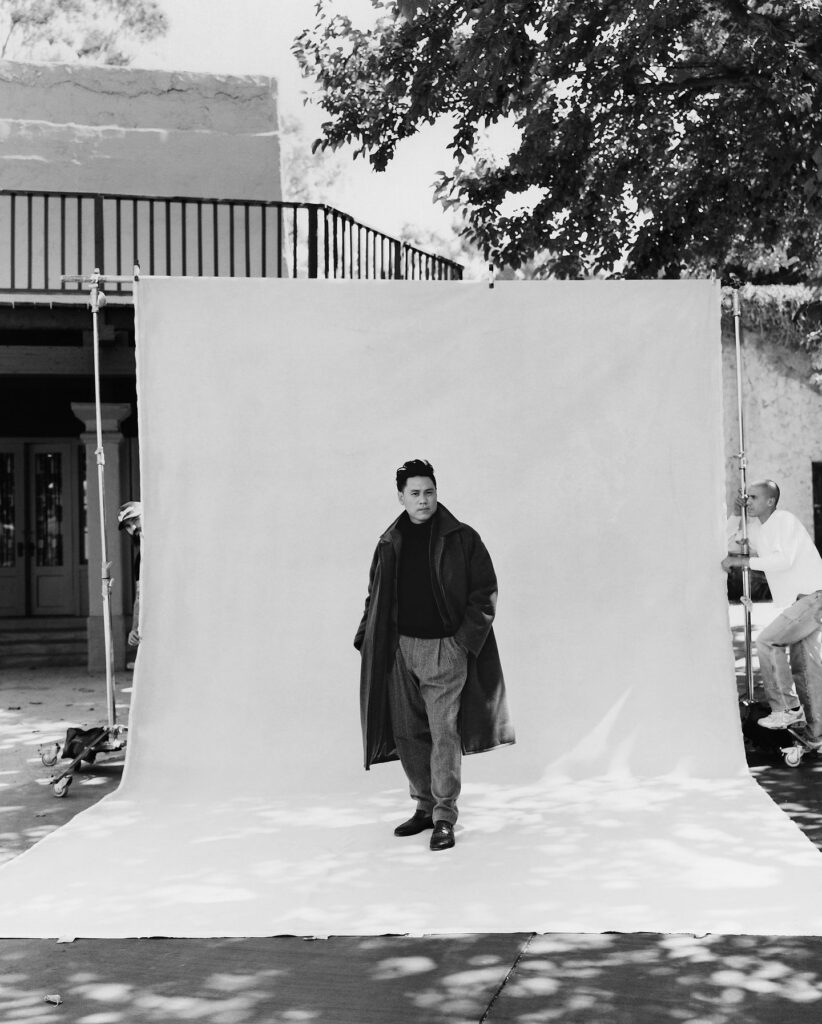 A black and white photo of a man standing on a light-colored backdrop, wearing a long coat and styled trousers, with trees casting dappled shadows in the foreground. A film crew member is visible in the background.