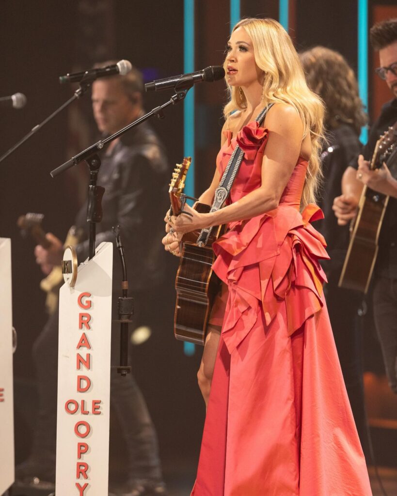 Carrie Underwood sings at the Grand Ole Opry, wearing a vibrant pink dress and holding an acoustic guitar, with band members performing in the background.