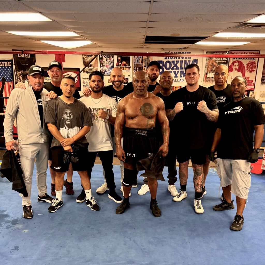 Mike Tyson stands shirtless at a boxing gym surrounded by ten trainers and supporters, all in athletic attire, showcasing teamwork and camaraderie as they prepare for an upcoming fight.