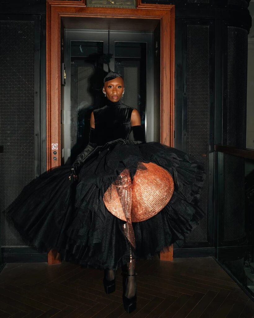 A woman in a dramatic black gown with a voluminous skirt and a striking metallic underskirt stands near an elevator. She wears long black gloves and has a sleek hairstyle.