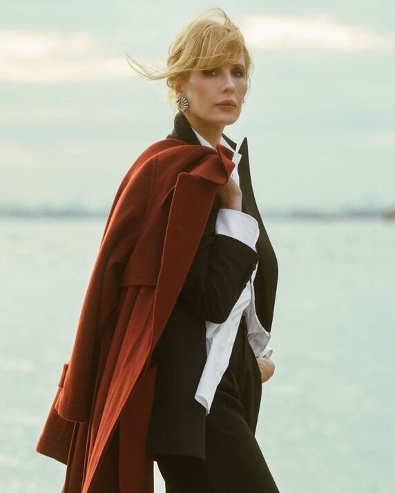 Kelly Reilly elegantly poses by the water, wearing a black outfit with a white shirt and a flowing red coat, exuding confidence and sophistication.