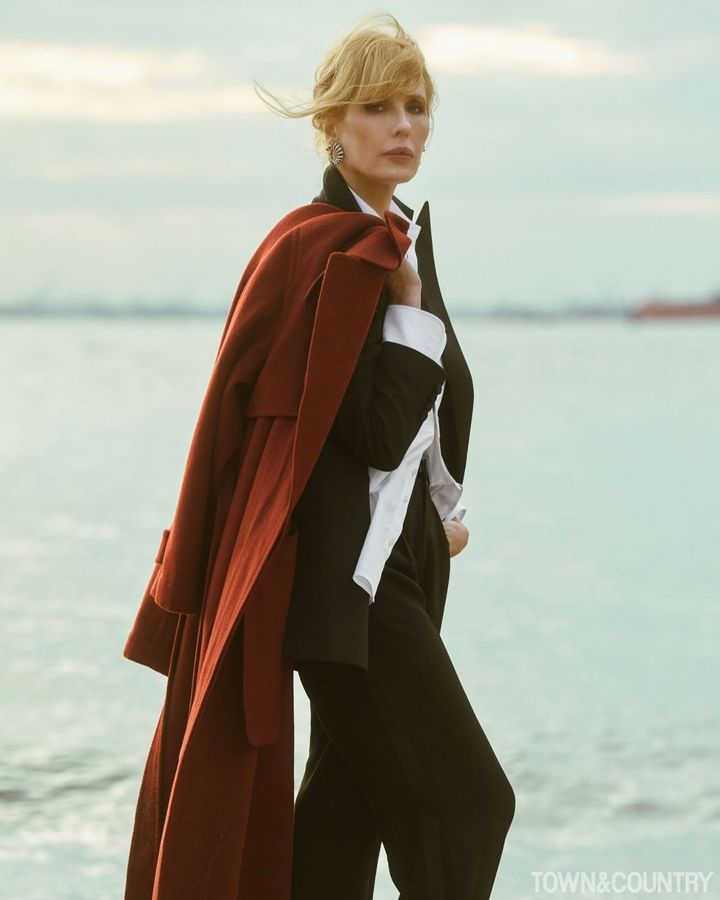 Kelly Reilly elegantly poses by the water, wearing a black outfit with a white shirt and a flowing red coat, exuding confidence and sophistication.