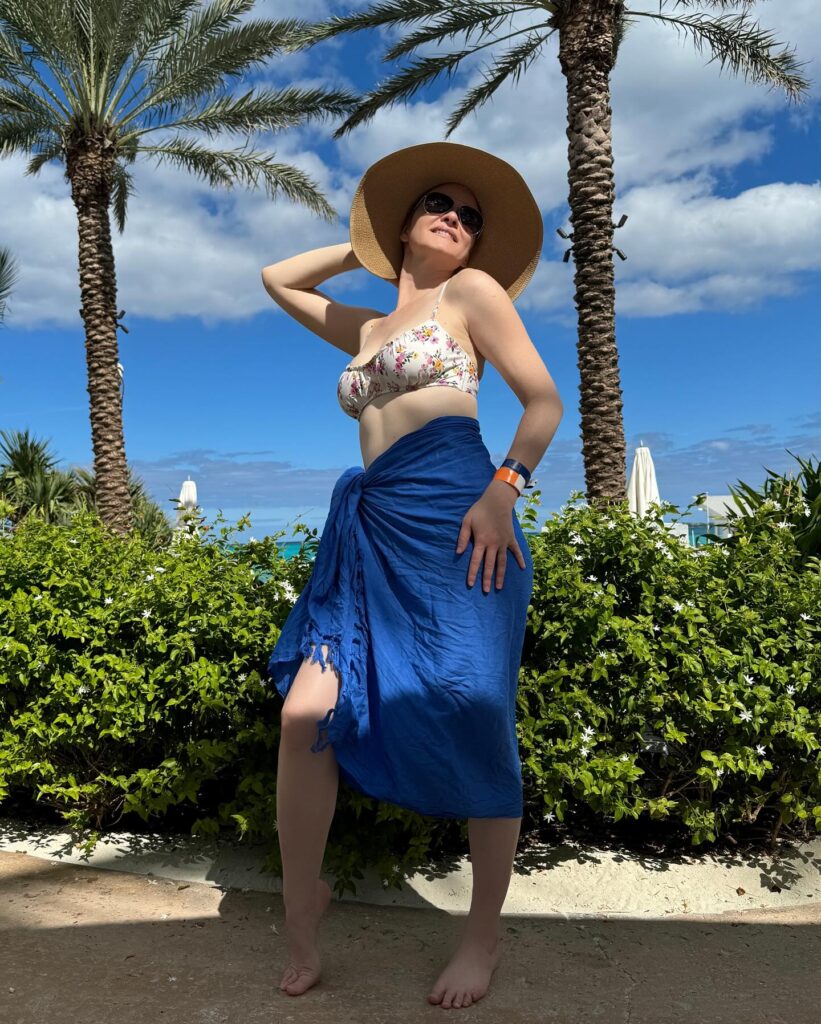 A woman poses outdoors in a floral bikini top and a blue sarong, wearing a wide-brimmed straw hat and sunglasses. She stands in front of lush greenery and palm trees, with a blue sky and clouds in the background.