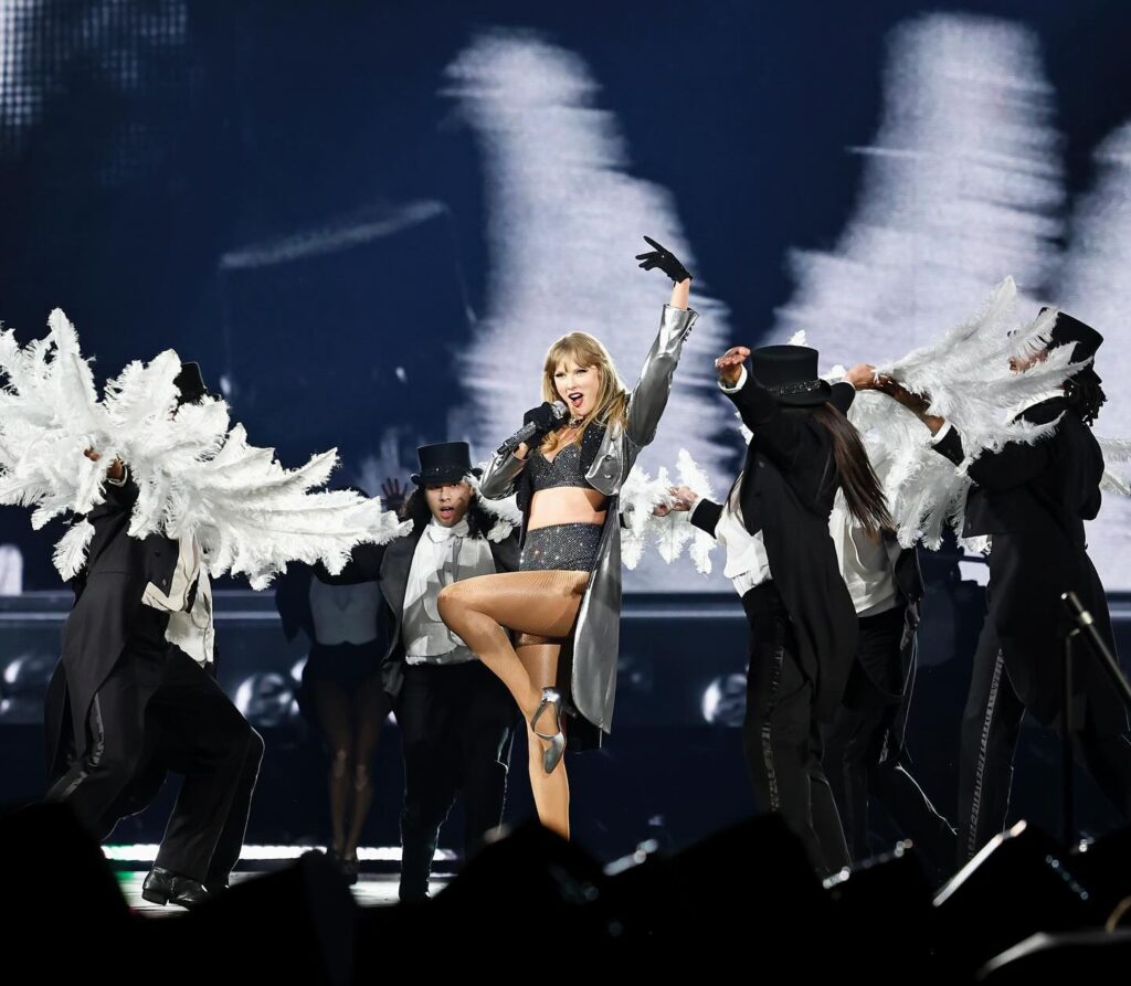  Taylor Swift performing on stage in a sparkling black outfit and silver jacket, surrounded by dancers in formal attire with feather props, striking a dynamic pose while singing passionately during her concert.