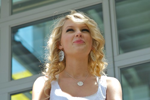 Taylor Swift smiling while wearing a white top and silver earrings, standing outdoors in front of large windows on a sunny day.