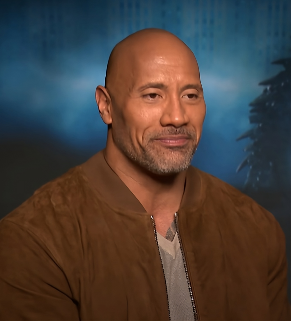 Dwayne Johnson smiling during an interview, wearing a brown jacket against a blurred blue background.