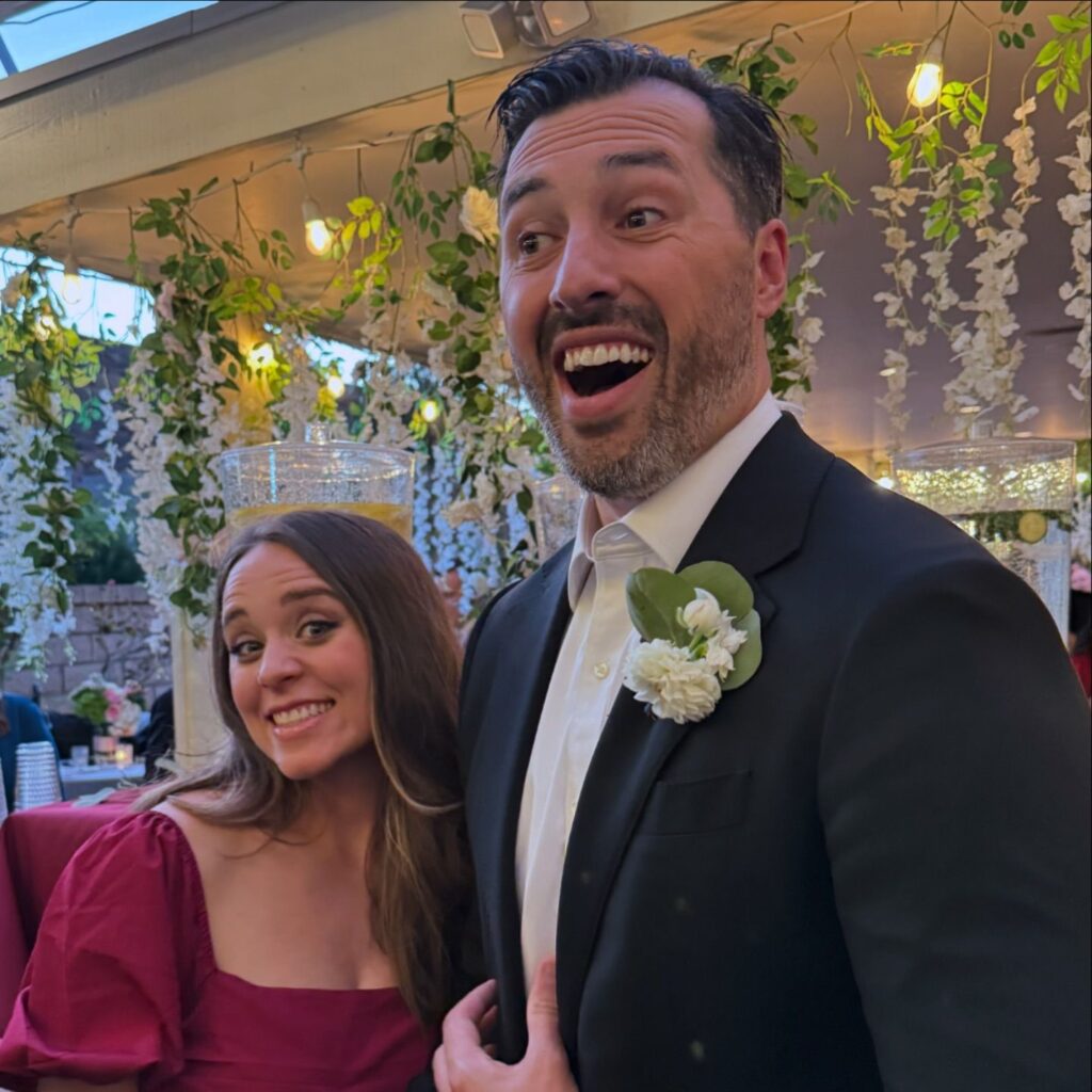 Jinger Duggar and Jeremy Vuolo joyfully pose together with wide smiles at a festive event, surrounded by elegant floral decorations and soft lighting. Jeremy is in a black suit with a floral boutonnière, while Jinger wears a burgundy dress.