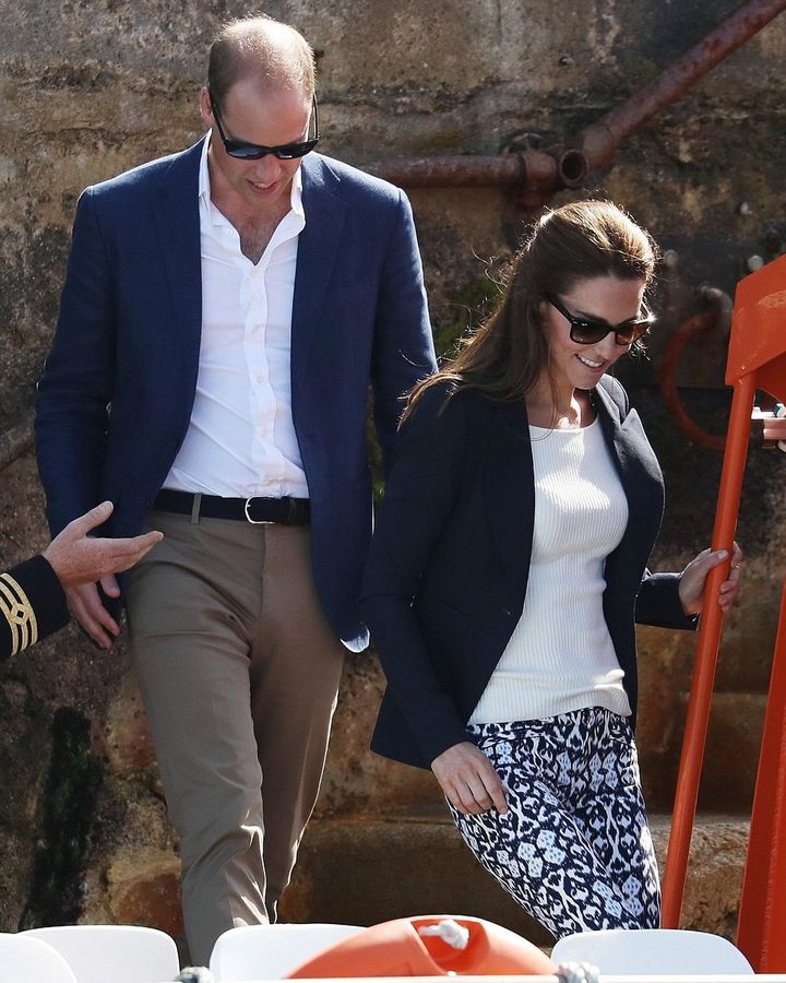  Prince William and Kate Middleton are walking together outdoors, both wearing sunglasses. William is dressed in a navy blazer over a white shirt, paired with beige trousers, while Kate is wearing a navy blazer over a white sweater and patterned pants. They appear to be enjoying a casual outing, with a rustic stone backdrop behind them.