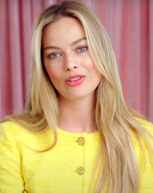 Margot Robbie, wearing a bright yellow jacket with a button detail, smiles warmly at the camera. She has long, flowing blonde hair and striking blue eyes, set against a soft pink background.