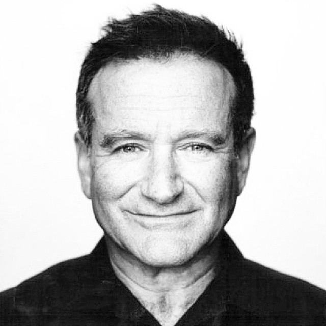 A black and white portrait of Robin Williams smiling warmly at the camera. He has short, dark hair and is wearing a black shirt, with a soft expression that reflects his approachable and charismatic personality.