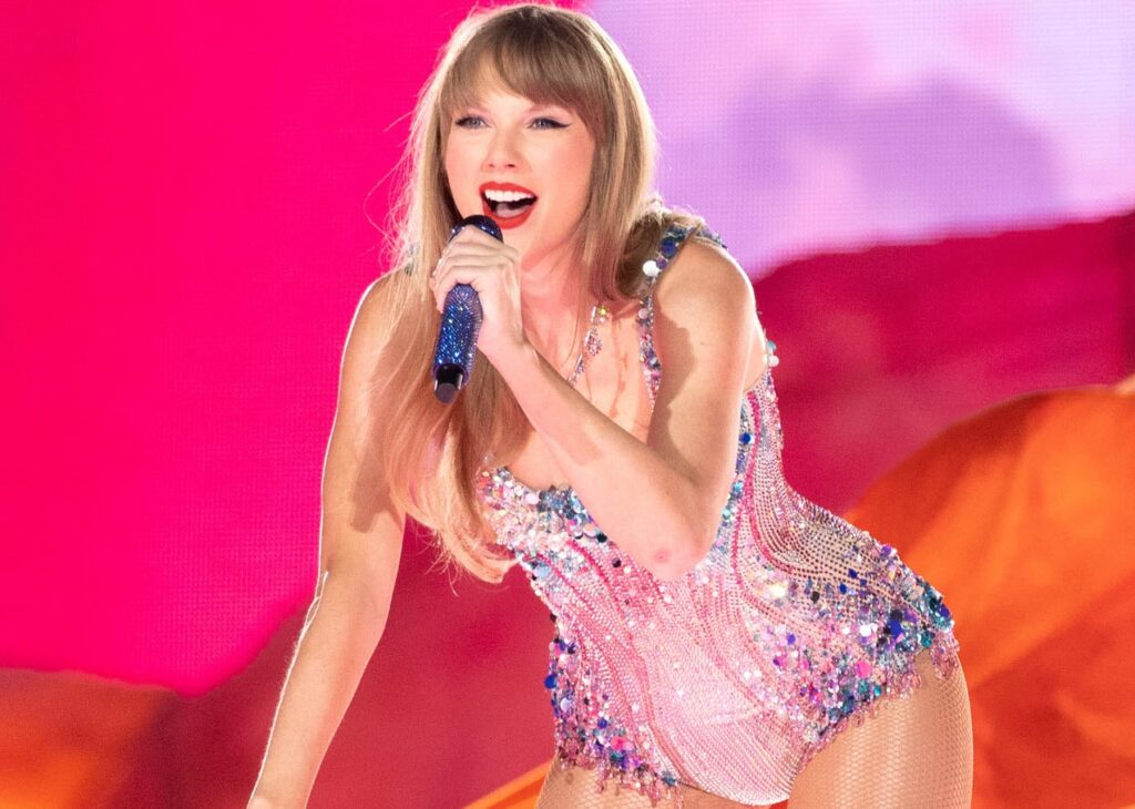 Taylor Swift performs passionately on stage wearing a sparkling costume, holding a microphone, with a vibrant pink background.