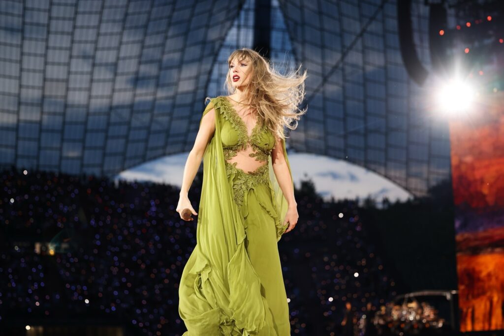 "Taylor Swift performs on stage wearing a flowing green dress with intricate detailing, her hair blowing in the wind, captivating the audience with her energy and presence under a beautifully lit sky."