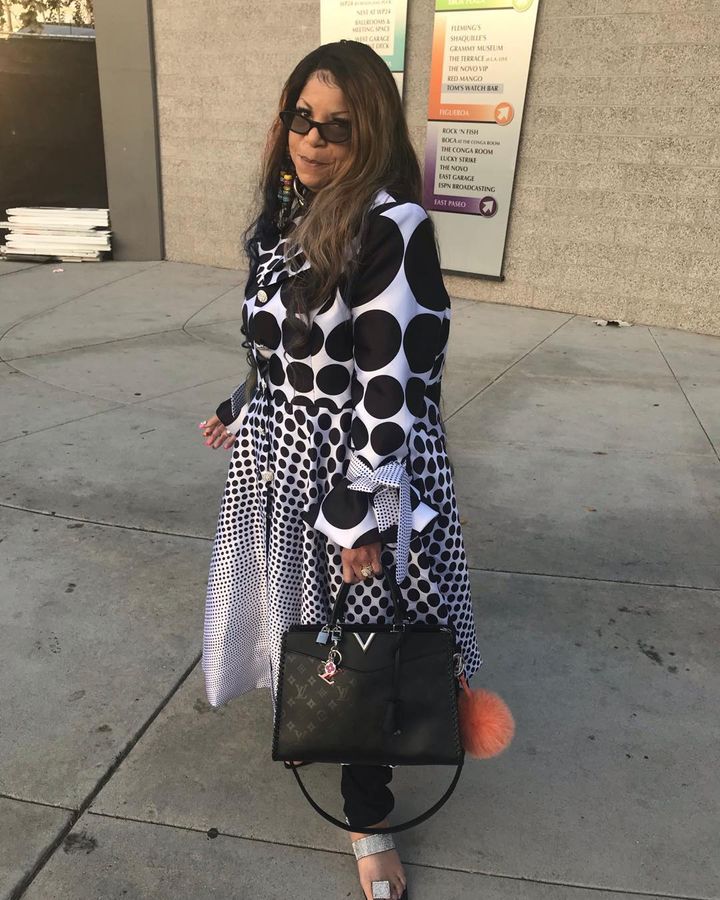 A woman wearing a polka dot dress and black sunglasses, holding a black handbag with a pink pom-pom, stands on a sidewalk.