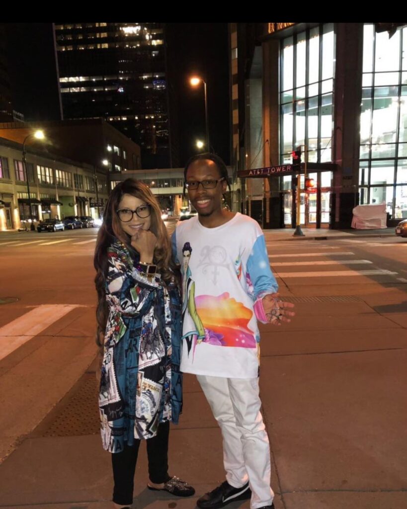 A woman and a man are posing for a photo on a city street at night. The woman has long, wavy hair and is wearing large glasses along with a colorful, patterned coat. The man is dressed in a white shirt featuring a vibrant graphic design, light pants, and black shoes. They are smiling and standing together in front of illuminated buildings.