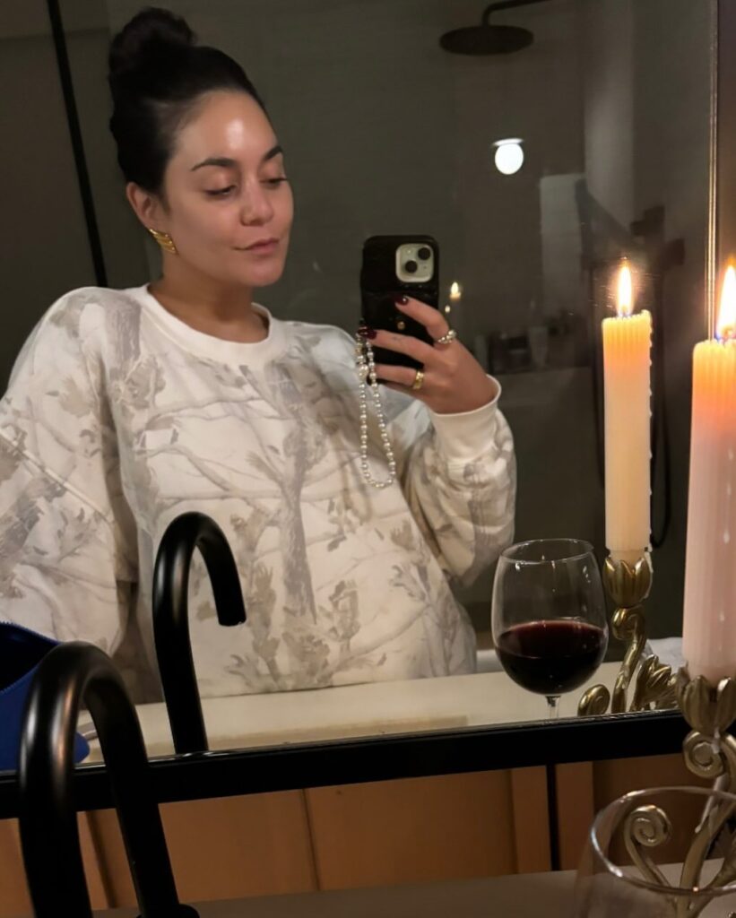  Vanessa Hudgens takes a mirror selfie wearing a cozy light-colored sweatshirt, showcasing her natural beauty with minimal makeup. She holds her phone in one hand and has a glass of red wine beside her on the table with flickering candles in the background.