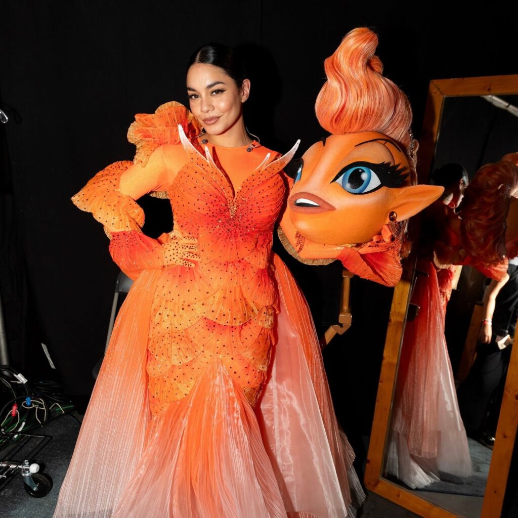 Vanessa Hudgens poses in an elaborate orange dress adorned with ruffles and sparkle, holding a whimsical mask of a character with a vibrant hairstyle.