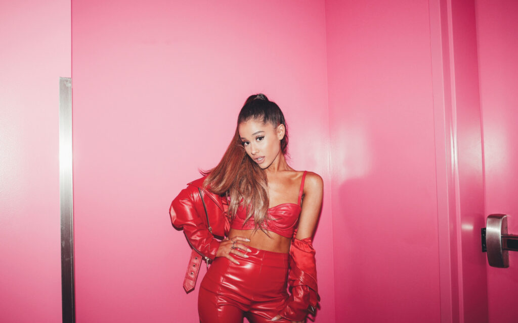 Ariana Grande poses confidently against a vibrant pink wall, wearing a matching red crop top and shiny pants, complemented by a stylish red jacket draped over her arm.