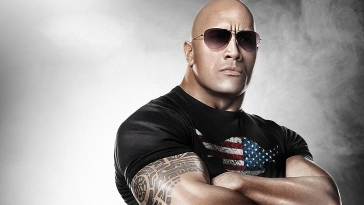 Dwayne Johnson, also known as The Rock, poses confidently with sunglasses and a black shirt featuring an American flag design, displaying his muscular build against a smoky background.