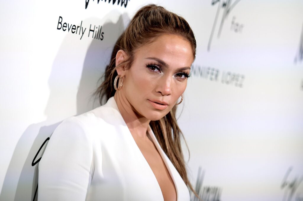 Jennifer Lopez poses at an event with a confident expression, wearing a deep-v white blazer and large hoop earrings against a minimalist backdrop.