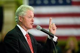 Bill Clinton speaking passionately at an event, gesturing with his finger, with a blurred American flag in the background.