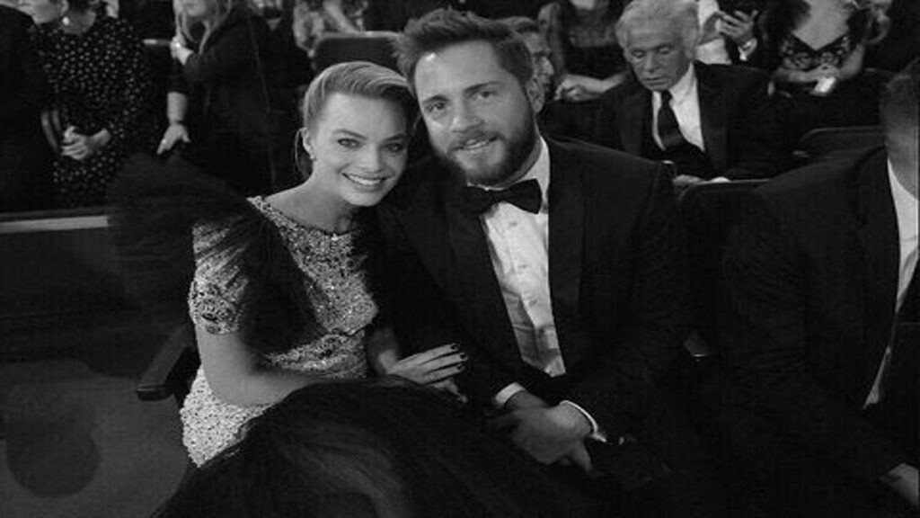 Margot Robbie and Tom Ackerley smiling together at an event, seated in formal attire.