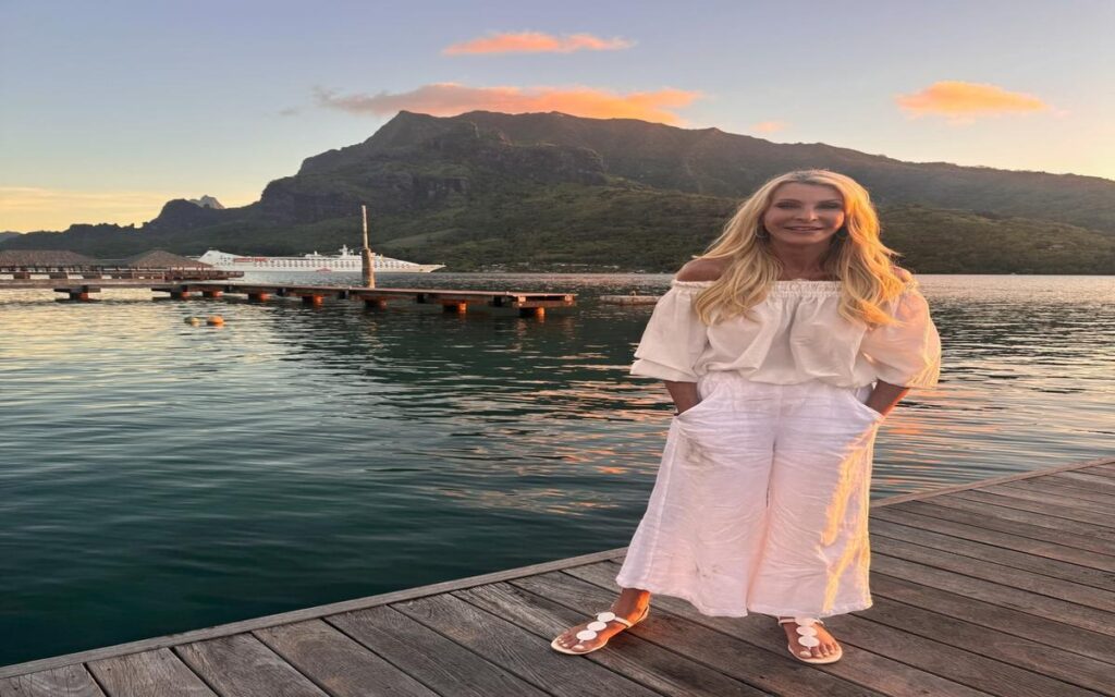 A woman in a white off-shoulder top and white linen pants stands on a wooden dock by a serene lake, with a mountainous backdrop during sunset.